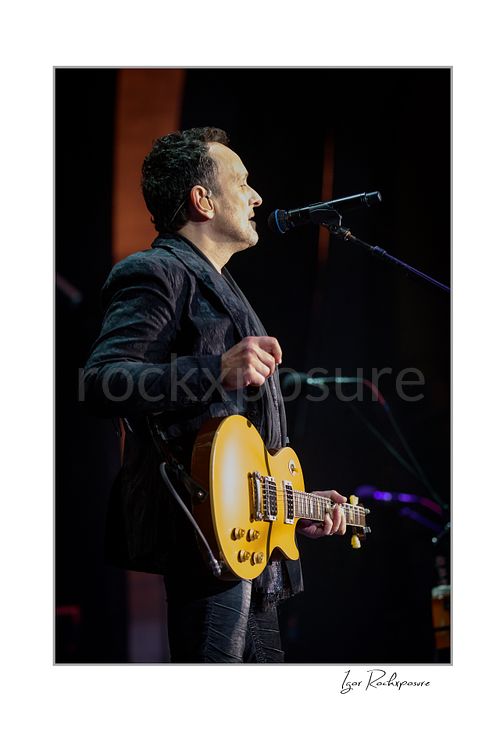 Vertical color image of Vivian Campbell of Def Leppard performing live on stage in profile, singing into a microphone while holding a gold electric guitar