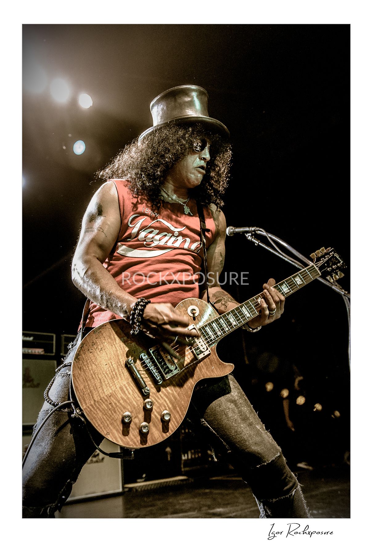 Vertical concert photography of Slash playing electric guitar wearing his signature top hat under warm stage lighting