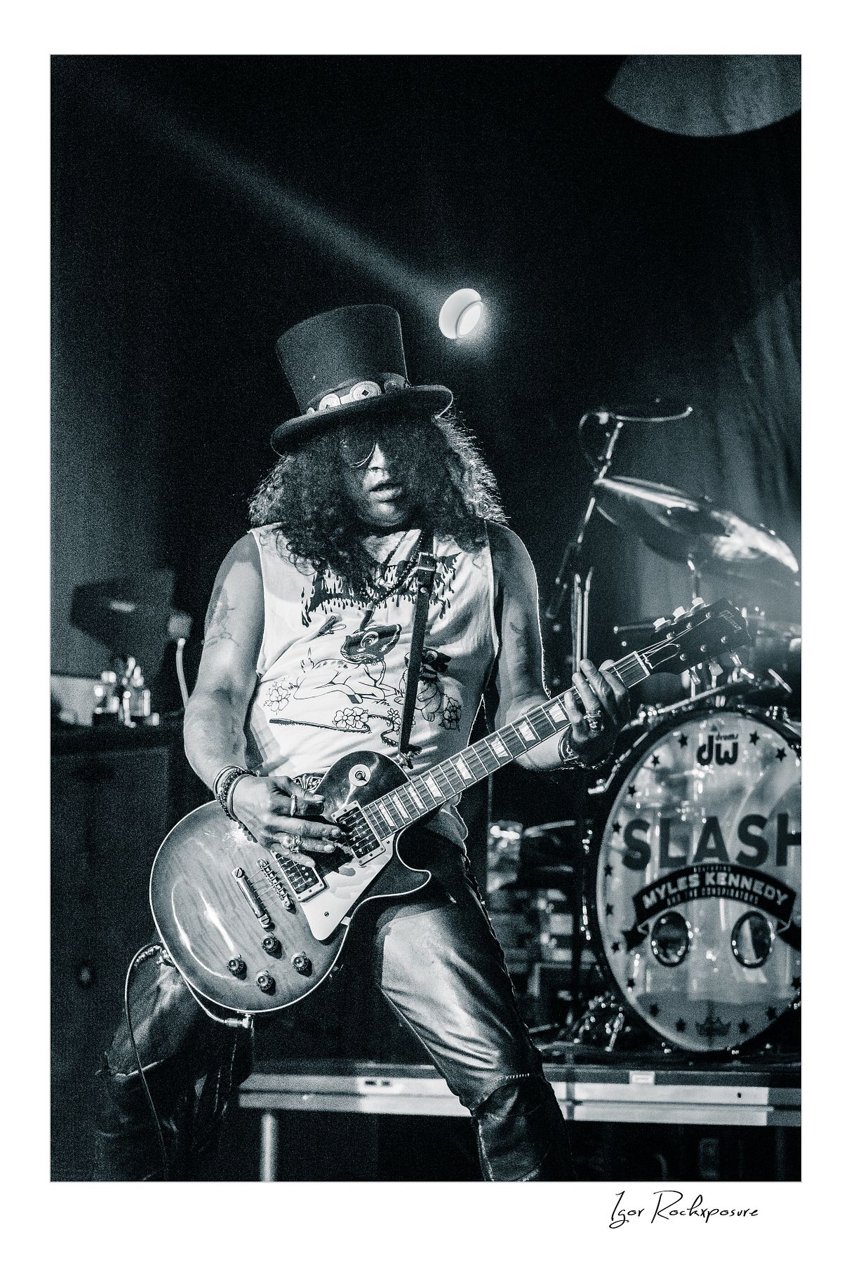 Vertical concert photography of Slash playing electric guitar wearing his signature top hat in black and white concert photography under dramatic stage lighting