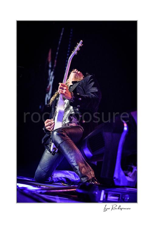 Vertical color low-angle image of Joel Hoekstra of Whitesnake performing live on stage, kneeling while playing electric guitar under purple concert lighting