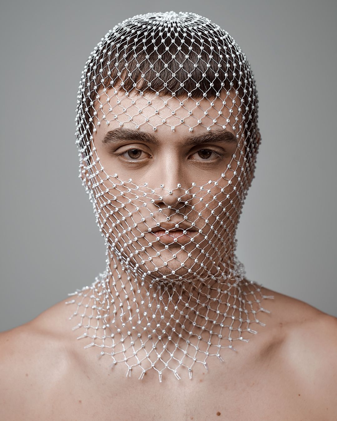 Editorial fashion portrait of a male model with short dark hair, styled in a silver beaded mesh headpiece covering his face and neck. Minimalist studio setup with soft lighting highlights the clean lines and futuristic design. Shot by a Nashville fashion and portrait photographer, ideal for men&rsquo;s editorial, branding, and creative campaigns.