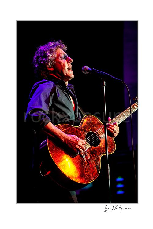 Vertical color image of Roger Daltrey singing into a microphone while playing guitar under vivid purple and warm concert light
