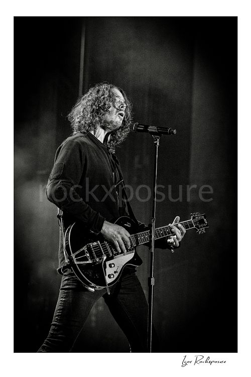 Vertical black and white image of Chris Cornell performing live on stage, playing electric guitar beside a microphone stand under concert lighting