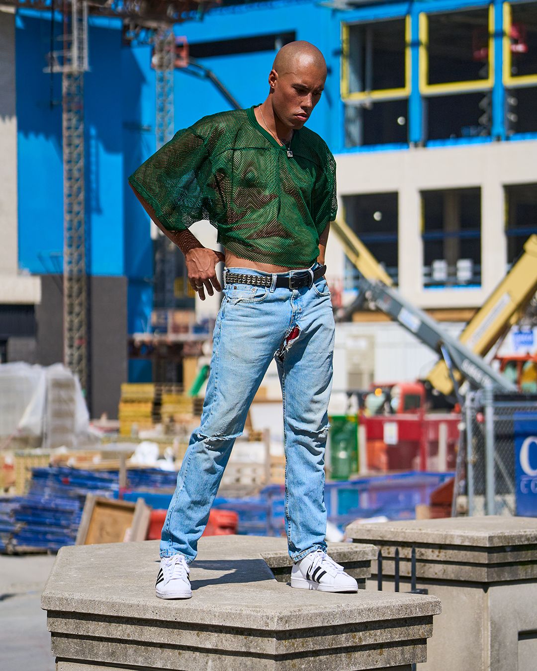 Male model wearing a green mesh shirt and light wash jeans, standing on top of a concrete pillar in front of a construction site in Nashville.