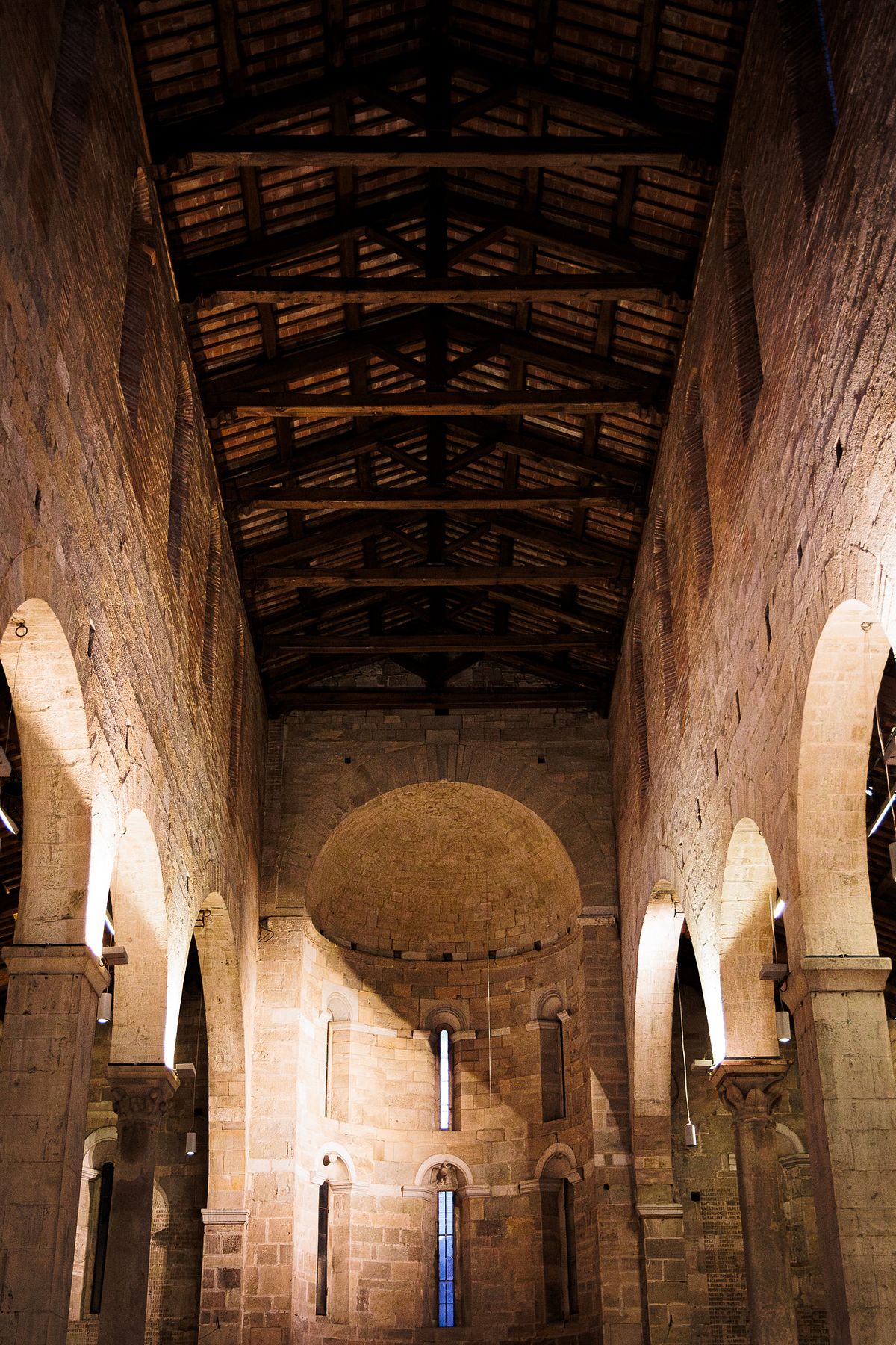 Interior view of a historic stone church in Lucca, Italy, showing the central nave with high wooden-beam ceiling, thick stone columns and tall arched windows on both sides, leading to a semi-circular apse at the far end, softly lit to highlight the warm textures of the stone and wood and creating a calm, serene atmosphere.
