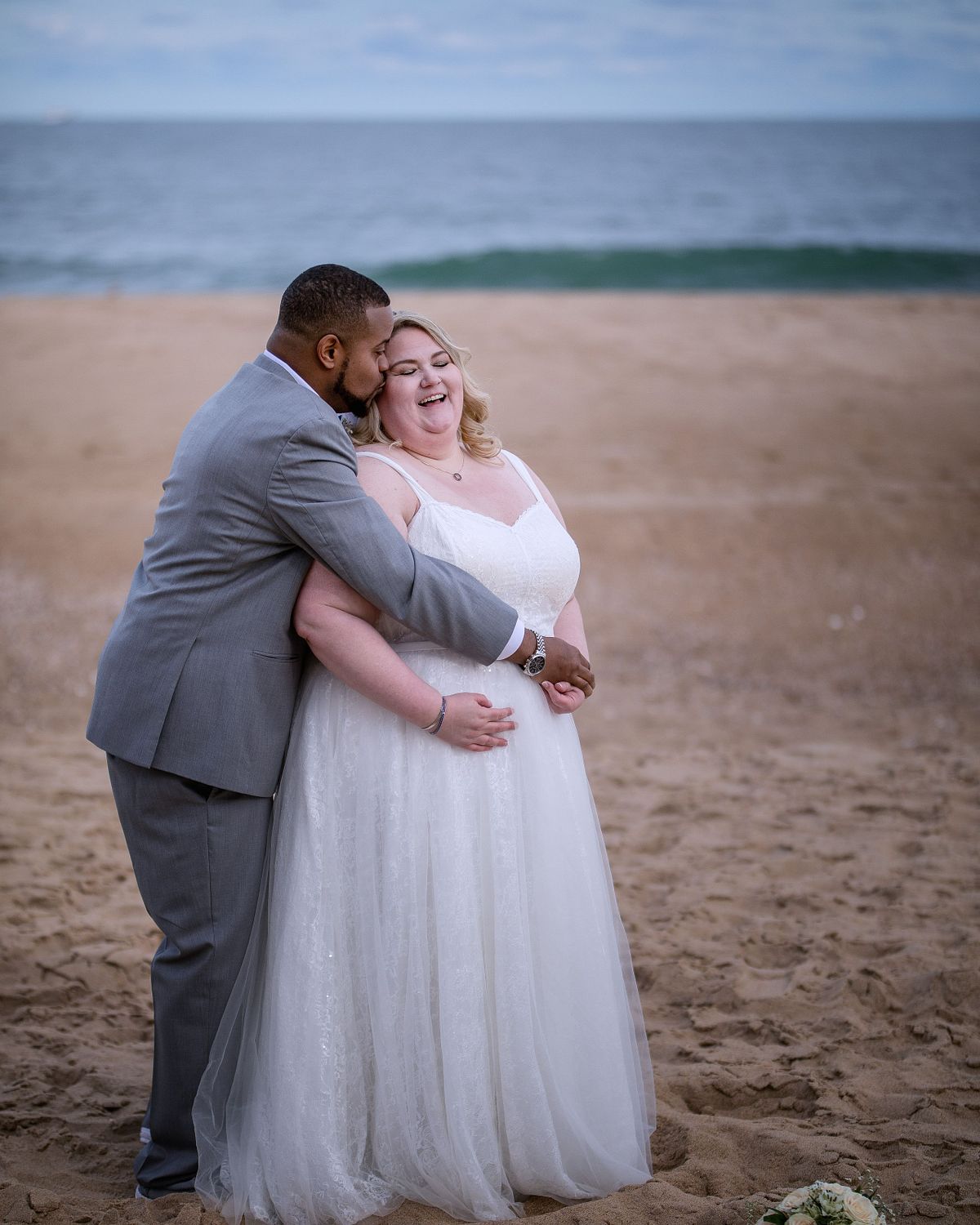 a bi-racial couple on the beach the groom is hugging his bride and she is smiling at him at Golden Sands ocean city, with ocean weaves behind them