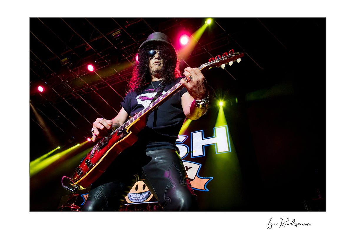 Horizontal concert photography of Slash playing electric guitar wearing his signature top hat under colorful stage lighting