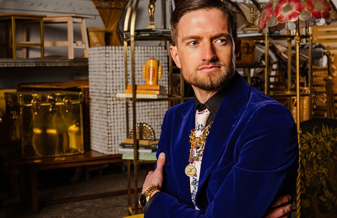 Brightly colored portrait of a male model wearing a royal blue velvet jacket inspired by a Gucci campaign photographed surrounded by furniture and antiques in a warehouse.
