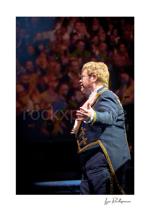 Vertical color image of Elton John on stage in profile with one arm extended before a blurred concert crowd