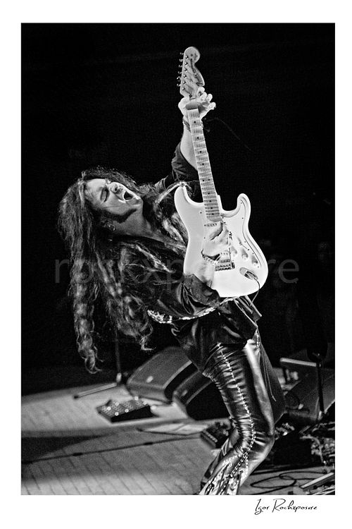 Vertical black and white image of Yngwie Malmsteen leaning back with a white guitar in a dramatic live performance pose, long hair flowing as he plays under dark stage light