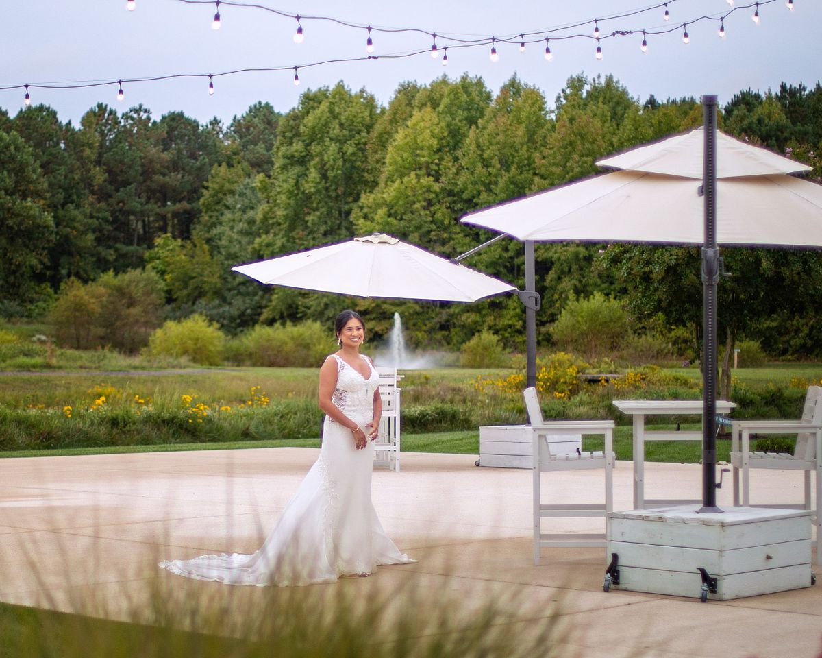 A bride in a lace gown smiling on an outdoor patio at Kylan Barn, showcasing soft natural light and a scenic fountain background.