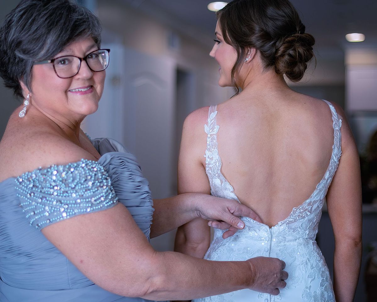 mom helping her daughter with the finishing touches on the wedding dress, the mom is wearing a blue dress and is smiling at the photographer at dewey beach, sussex county, de