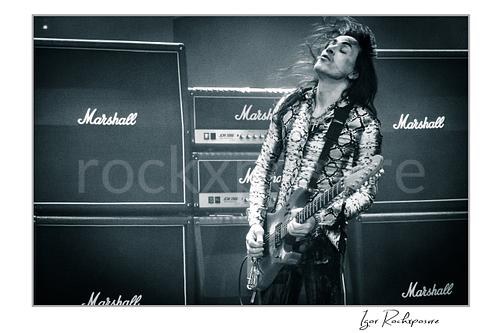 Horizontal black and white image of Nuno Bettencourt performing live on stage with an electric guitar, windblown hair, and a wall of Marshall amplifiers behind him