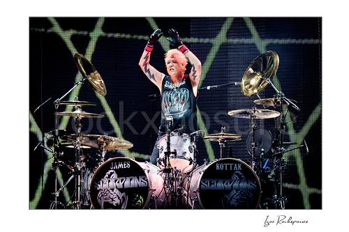 Horizontal color image of James Kottak of Scorpions behind the drums with raised arms, black gloves, and full Scorpions drum setup