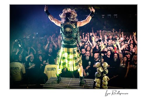 Wide shot from behind Zakk Wylde on stage with arms raised, wearing a kilt and vest, cheering the large crowd.