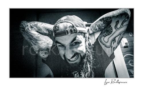 Horizontal black and white image of Mike Portnoy grinning at the camera with tattooed arms raised behind his head and a backward cap