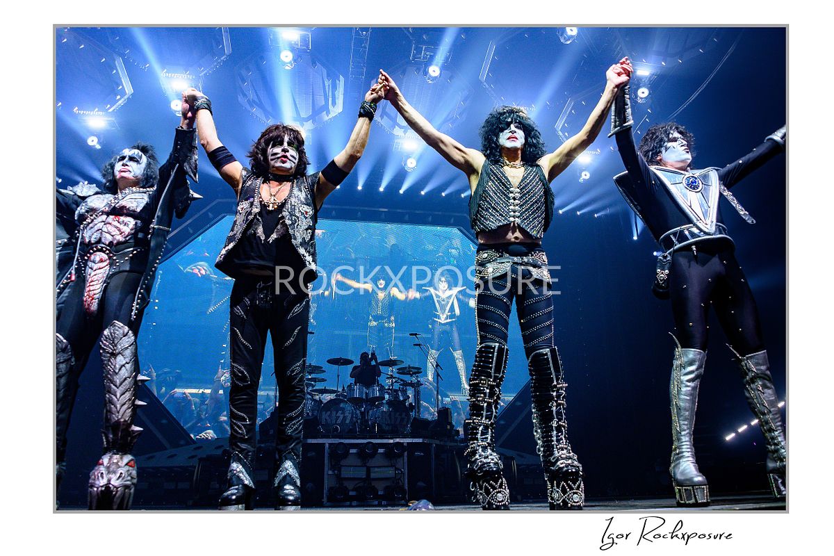 Horizontal color photograph of the full KISS band in full makeup and costumes standing with their backs to the stage and their hands raised toward the crowd as bright concert lighting fills the finale moment
