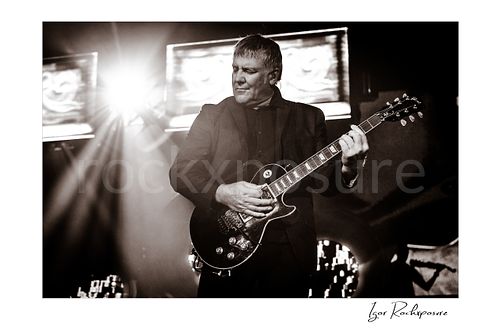 Horizontal black and white image of Alex Lifeson performing with electric guitar during a Rush concert with bright stage flare behind him