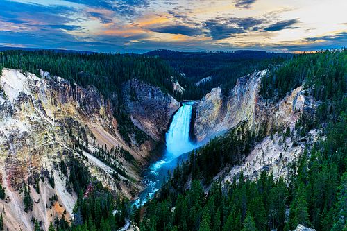 A horizontal view of the Lower Falls of the Yellowstone River plunging into the Grand Canyon of the Yellowstone at sunset, surrounded by steep canyon walls and evergreen forests.