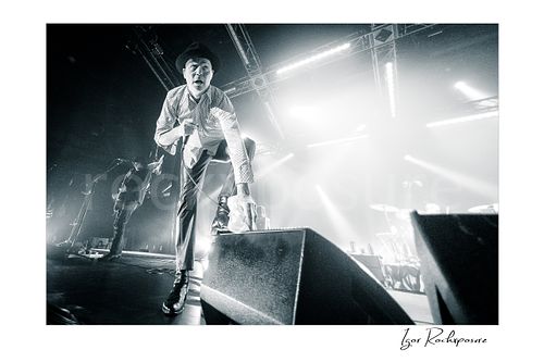 Horizontal black and white image of Gord Downie of The Tragically Hip in a dramatic low-angle wide-lens live performance shot, leaning toward the front of the stage with microphone and white cloth in hand under intense concert light