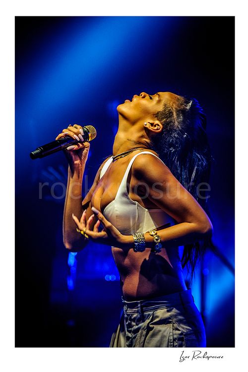 Vertical color image of Rihanna holding a microphone with head tilted back under deep blue concert lighting