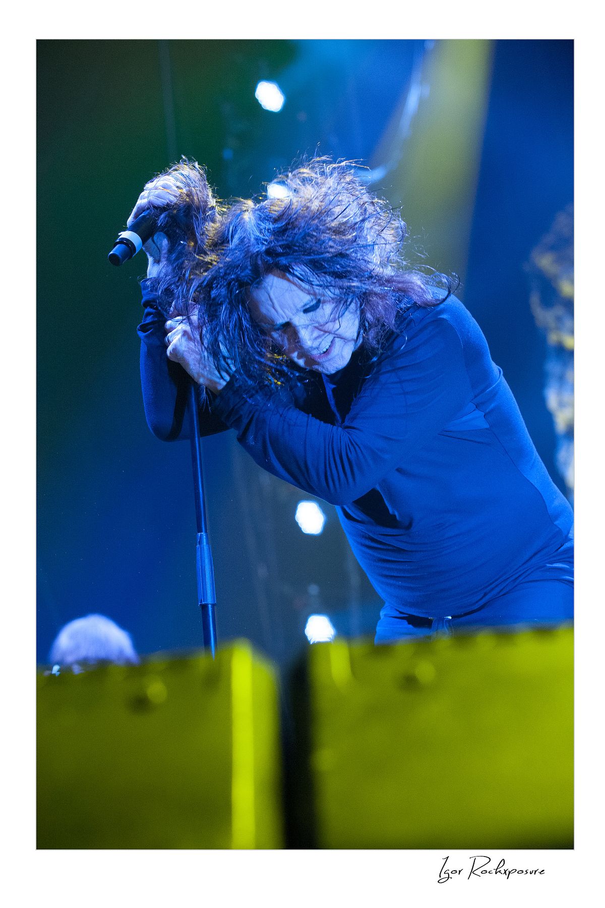 Ozzy Osbourne of Black Sabbath leaning forward singing into a microphone with intense expression under blue and yellow concert lighting
