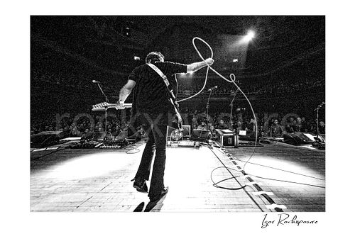 Horizontal black and white image of Steve Vai on stage from behind with guitar and a looping cable before a theatre audience