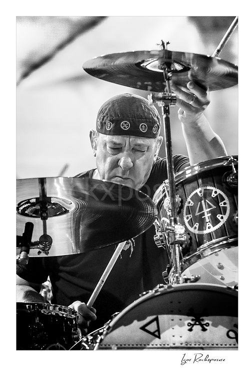 Vertical black and white image of Neil Peart performing behind his drum kit during a Rush concert, partially framed by cymbals with intense focus