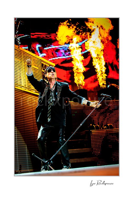 Vertical color image of Klaus Meine of Scorpions performing live on stage with microphone stand, sunglasses, and flames rising behind him