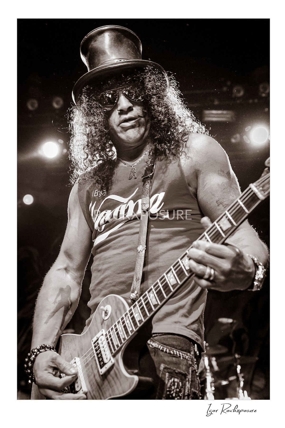 Vertical concert photography of Slash playing electric guitar wearing his signature top hat in black and white photography under stage lighting