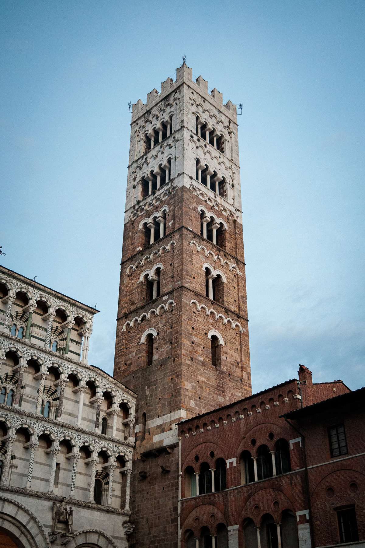 View of a tall historic stone bell tower in Lucca, Italy, standing against a blue sky, flanked on the left by a light stone building with dark stripes, arches and columns in a Romanesque style, and on the right by a red brick structure with rounded arches, all softly lit to highlight their architectural details.