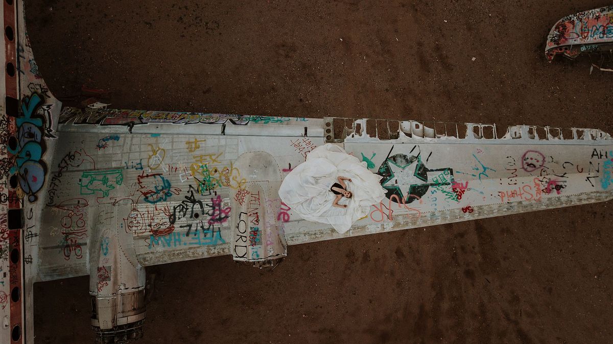 Striking aerial boudoir image of a woman laying across the wing of an abandoned airplane, wearing a flowing parachute skirt, captured by drone against the Arizona desert backdrop.