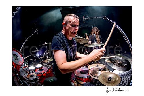 Horizontal color image of Korn drummer Ray Luzier at the drums with a drumstick raised, a scary expression, and bright stage lighting