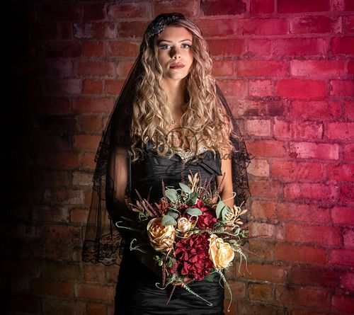 Model wearing a black wedding dress from Bridal Re-Loved Leicester, looking forward with a pink backdrop on a brick wall, photographed by Weddings by Jermaine, with hair and makeup by Justaboutweddingshair and beauty, and accessories by Sydney Grace Bridal