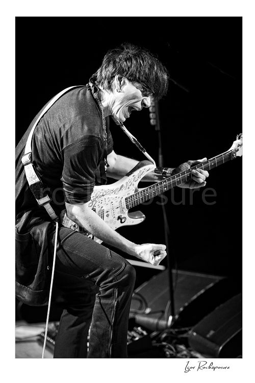 Vertical black and white image of Steve Vai playing guitar with an intense expression and clenched fist in a dramatic stage pose