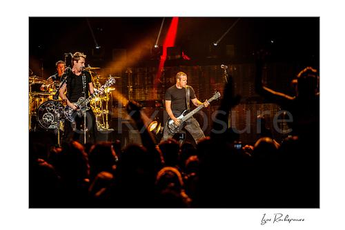 Horizontal color image of Chad Kroeger and Mike Kroeger of Nickelback performing onstage with the drummer visible behind them and a silhouetted crowd in front