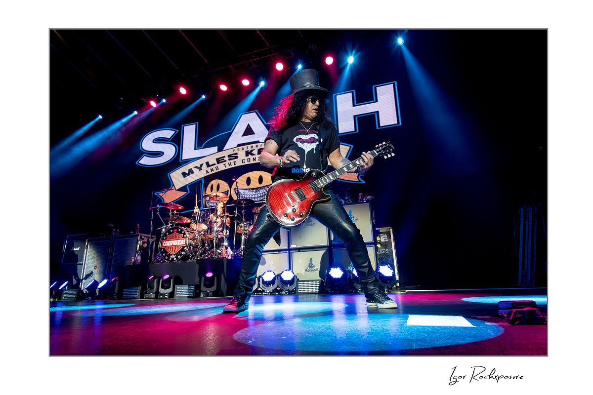 Horizontal concert photography of Slash playing electric guitar in a wide stance under colorful stage lighting with Slash stage backdrop