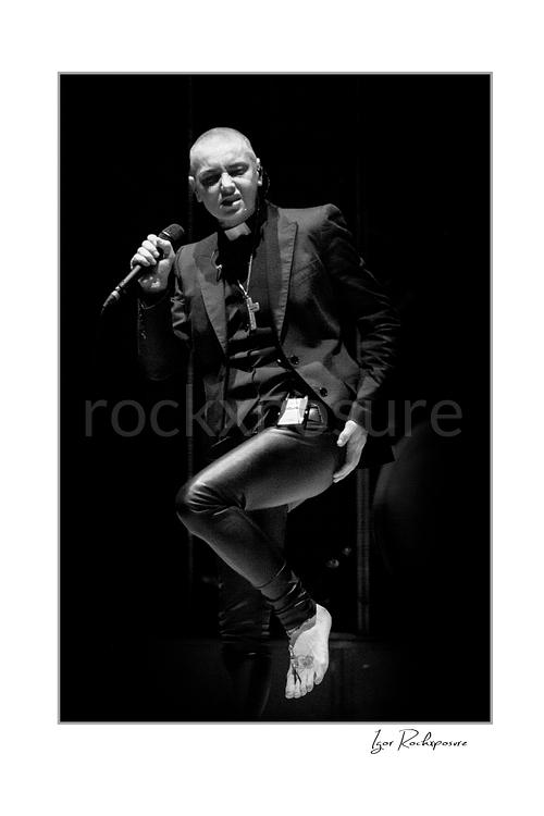 Vertical black and white image of Sinéad O’Connor holding a microphone on stage in a dramatic low-light portrait