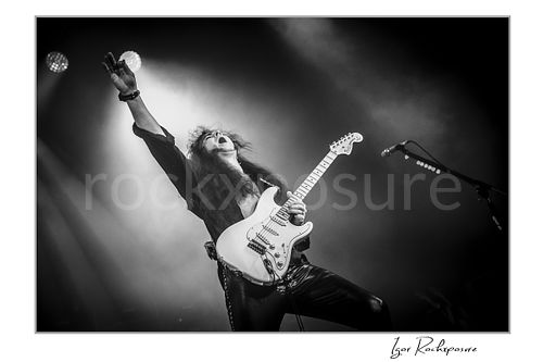Horizontal black and white image of Yngwie Malmsteen performing live with a white guitar, one arm raised high in a dramatic stage pose under bright backlight and concert haze