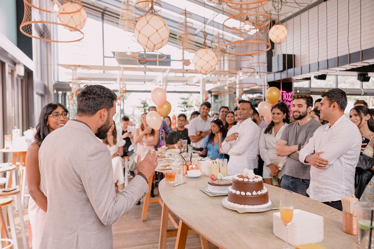 Photographer in Basel capturing a man at his engagement party in a suit speaking to a crowd at his celebration with his fiance. Attendees smile, surrounded by balloons and cake, in a bright, festive setting.