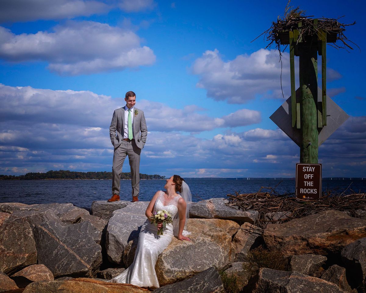 bride and groom posing  on a jetty, the groom is standing looking down at the bride, while the bride is sitting looking up at the groom