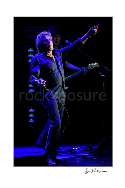 Vertical color image of Roger Daltrey performing onstage with one arm raised and microphone cable in hand under deep blue concert light