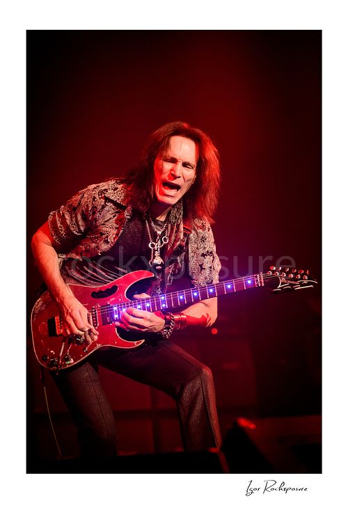 Vertical color image of Steve Vai playing guitar under vivid red stage lighting with glowing fret markers and an intense expression