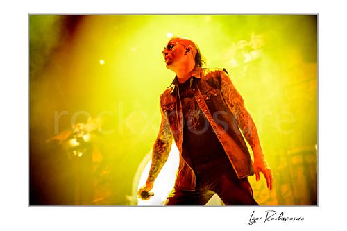 Horizontal color image of M. Shadows of Avenged Sevenfold on stage in a dramatic close-up with tattooed arms, sunglasses, and vivid yellow light