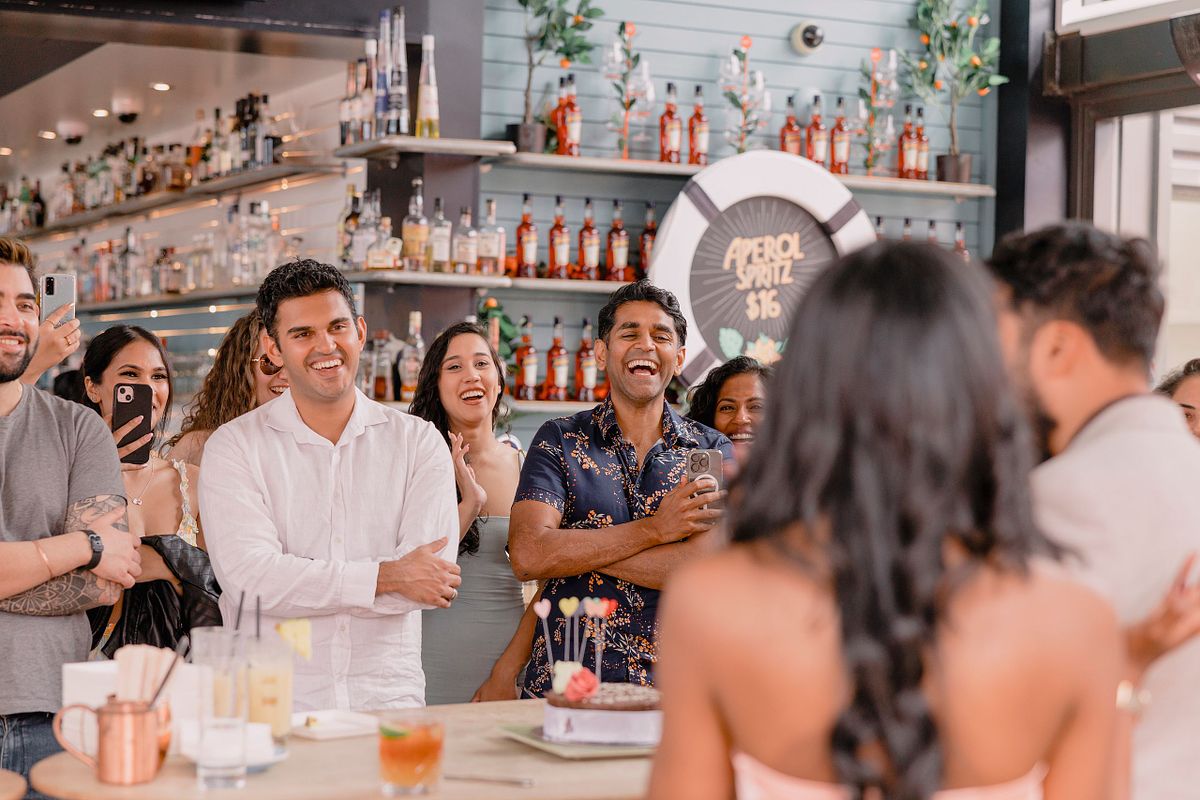 Photographer in Basel capturing a group of people at a lively engagement party, smiling and laughing, focus on a woman with long hair in the foreground. Bottles line the shelves, creating a festive atmosphere.