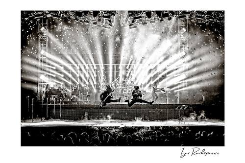 Horizontal black and white image of Trans Siberian Orchestra performing on stage with two jumping guitarists, dramatic light beams, and a packed concert crowd