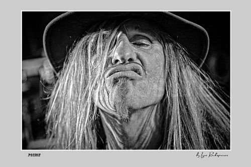 Horizontal black and white image of C.C. DeVille in an extreme close-up live portrait with long hair, hat and exaggerated expression