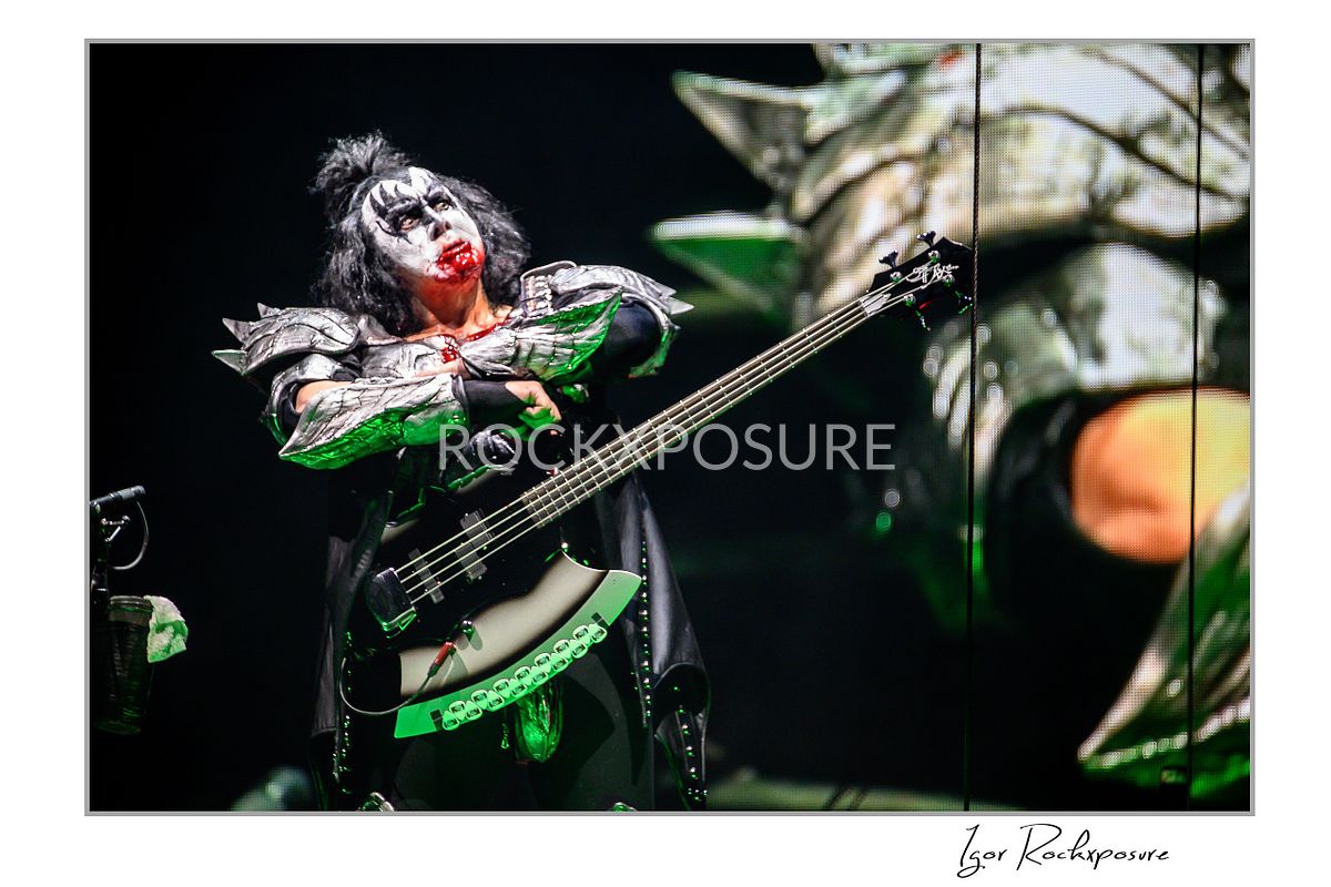 Gene Simmons of KISS in demon makeup with blood covering his mouth looking upward under green concert stage lighting during a dramatic stage moment