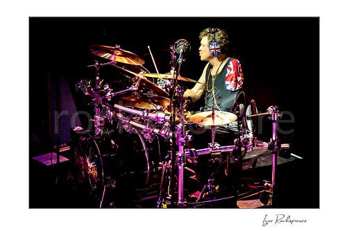 Horizontal color image of Rick Allen of Def Leppard performing live on stage behind his drum kit, wearing Union Jack headphones under bright concert lighting
