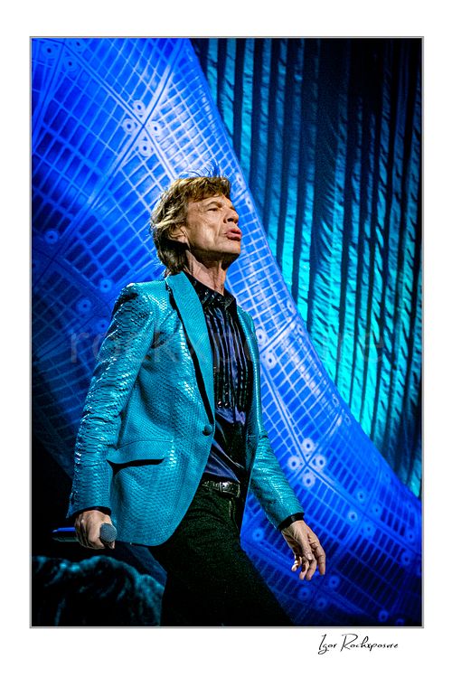 Vertical color image of Mick Jagger of The Rolling Stones walking on stage in a bright blue jacket against a vivid blue concert backdrop